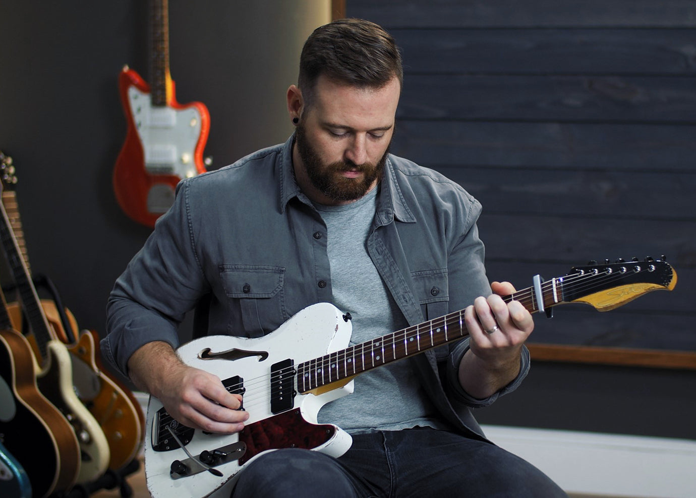 Man playing a white electric guitar in a room with guitars on the wall.