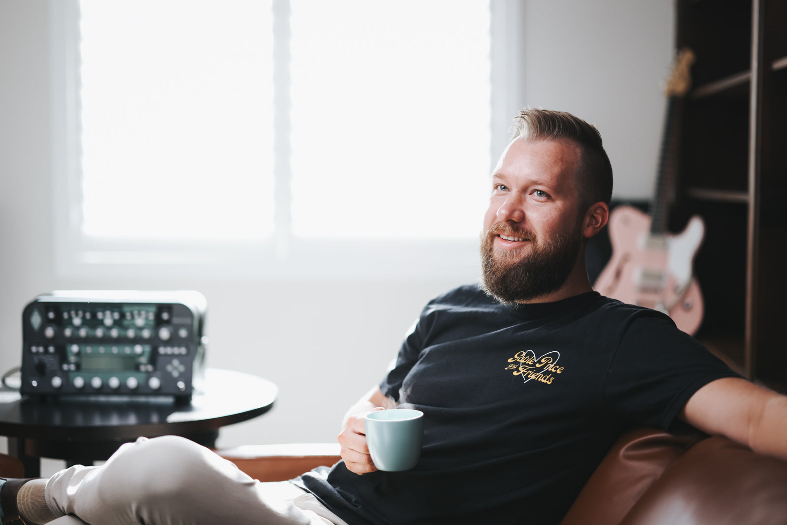 Man sitting on a couch holding a mug, with a guitar amplifier and window in the background